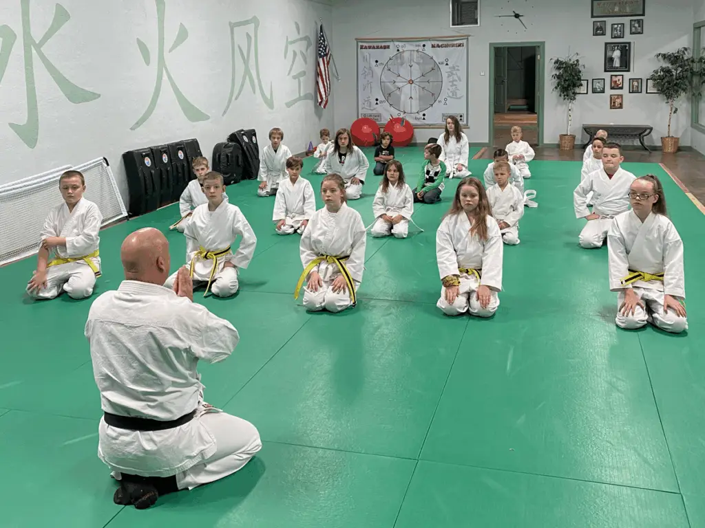 Children in karate uniforms sitting respectfully with their instructor at Bushido of Benson dojo, demonstrating focus and discipline.
